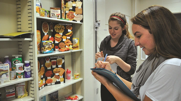 Two students working for their dietetics degrees stand in front of shelves of colorful displays of labeled food information.